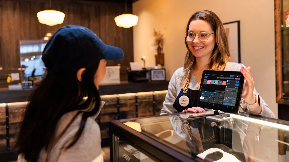 A shopkeeper serving a customer at the counter, ringing the sale up on BelloPOS running on a tablet